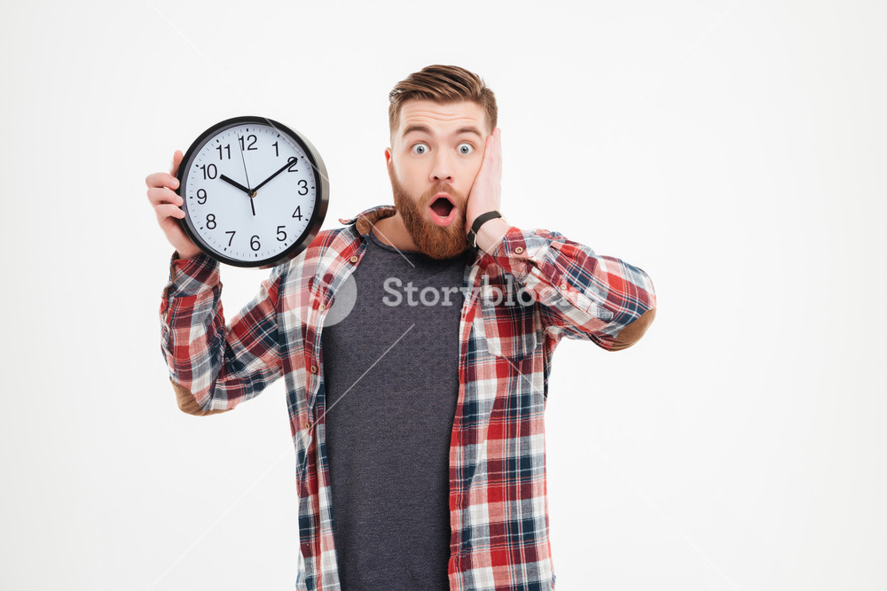 Surprised young man in plaid shirt holding wall clock over white background