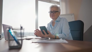 Mature gray hair woman smiling, reading message on mobile phone typing answer, chatting friends, relatives or husband on workplace in office