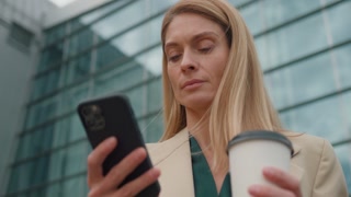 Caucasian Successful Woman uses Smartphone and holding a Cup of Coffee. Confident Middle-Aged Woman in Formal Wear, Taking a Break, Browsing on Social Media