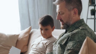 Tilt up shot of bearded male army veteran in uniform sitting on couch and showing photos to his son while sharing deployment stories